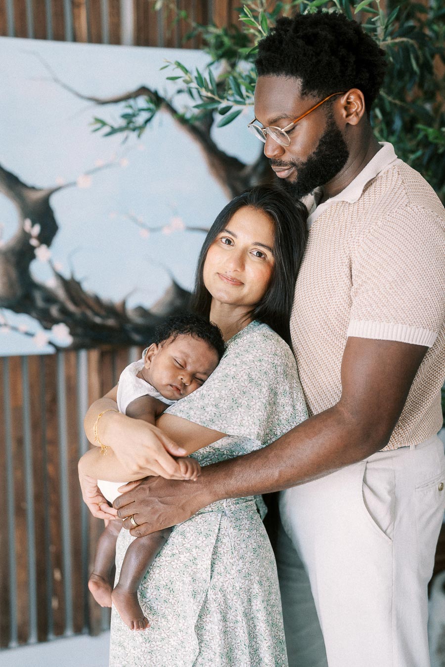 Karina holds sleeping Isabella while Terence embraces them beside a painted tree mural.