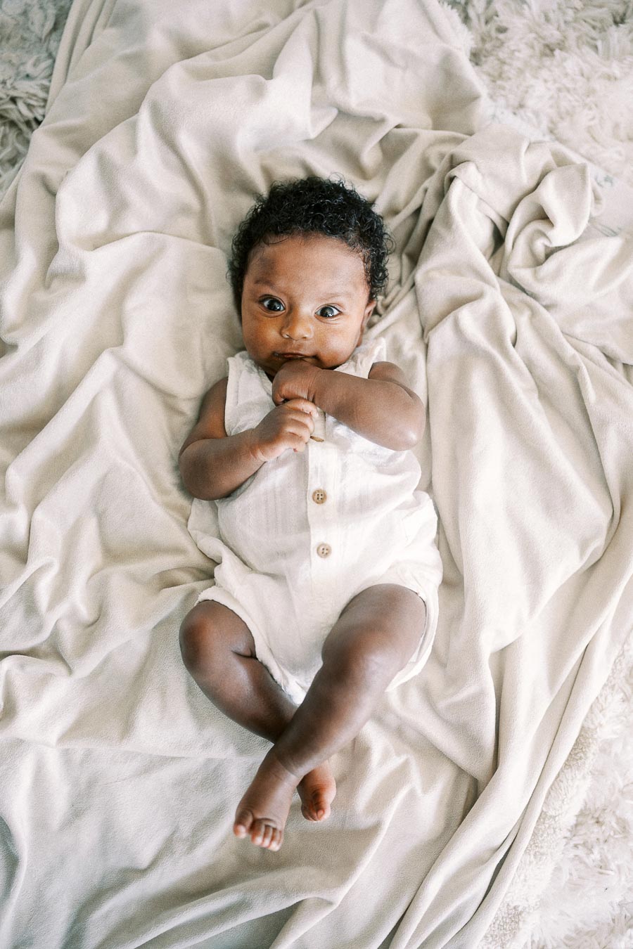 Baby Isabella lying awake on soft white blankets in a sleeveless romper.