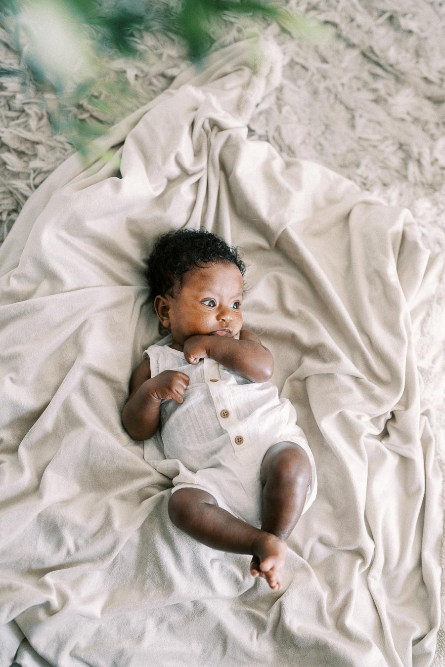 Baby Isabella lying on cream blankets in a white romper, looking to the side.