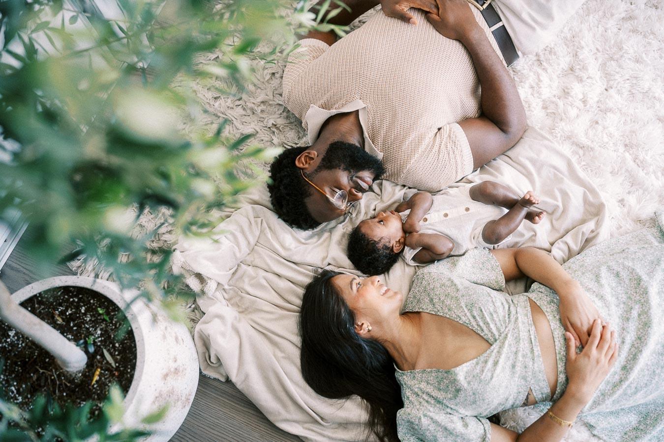 Parents lying on a blanket with their baby daughter beside a leafy indoor plant.
