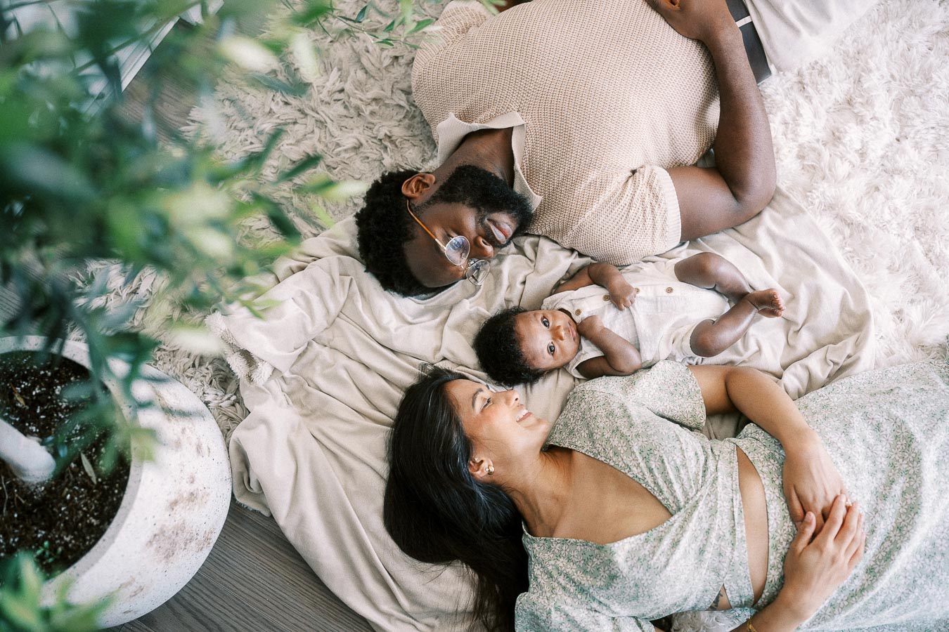 Karina and Terence lie beside newborn Isabella on a soft bed near a leafy plant.