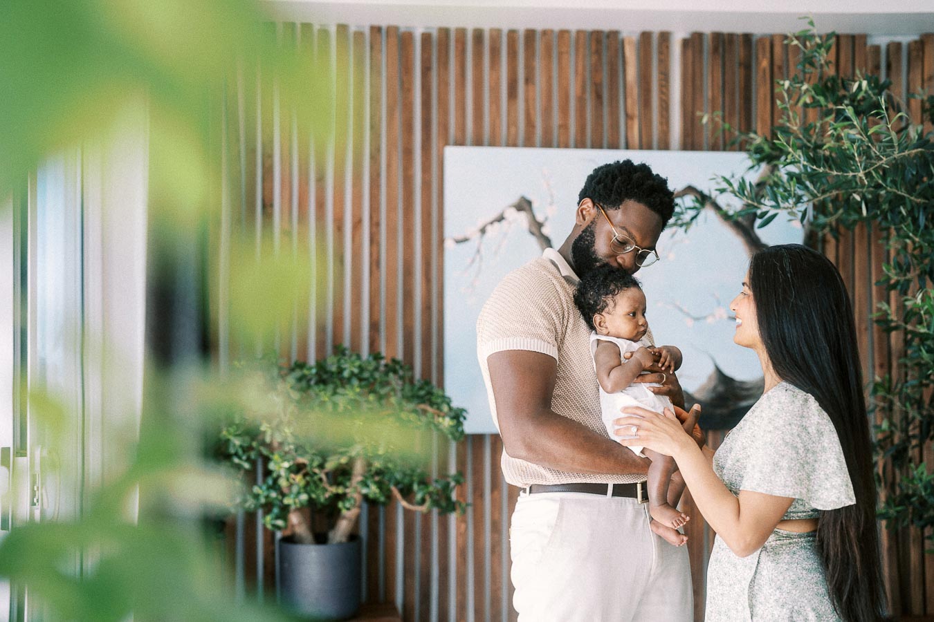 Karina and Terence holding Isabella beside indoor plants and a wood slat wall