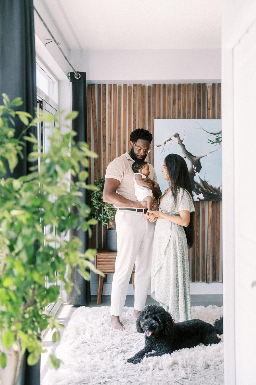 Parents hold newborn Isabella beside bright windows while their black dog rests on a white rug.