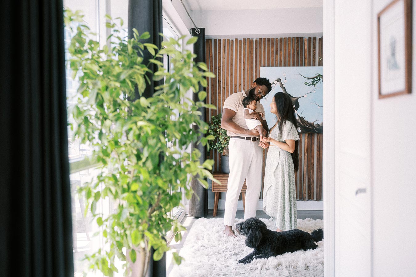 Parents holding newborn daughter by bright windows as a black dog rests on a white rug.