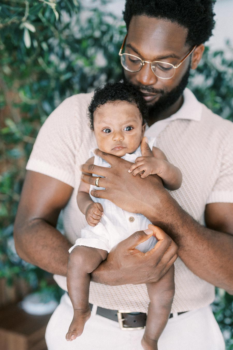 Terence cradles baby Isabella against his chest near leafy plants at home.