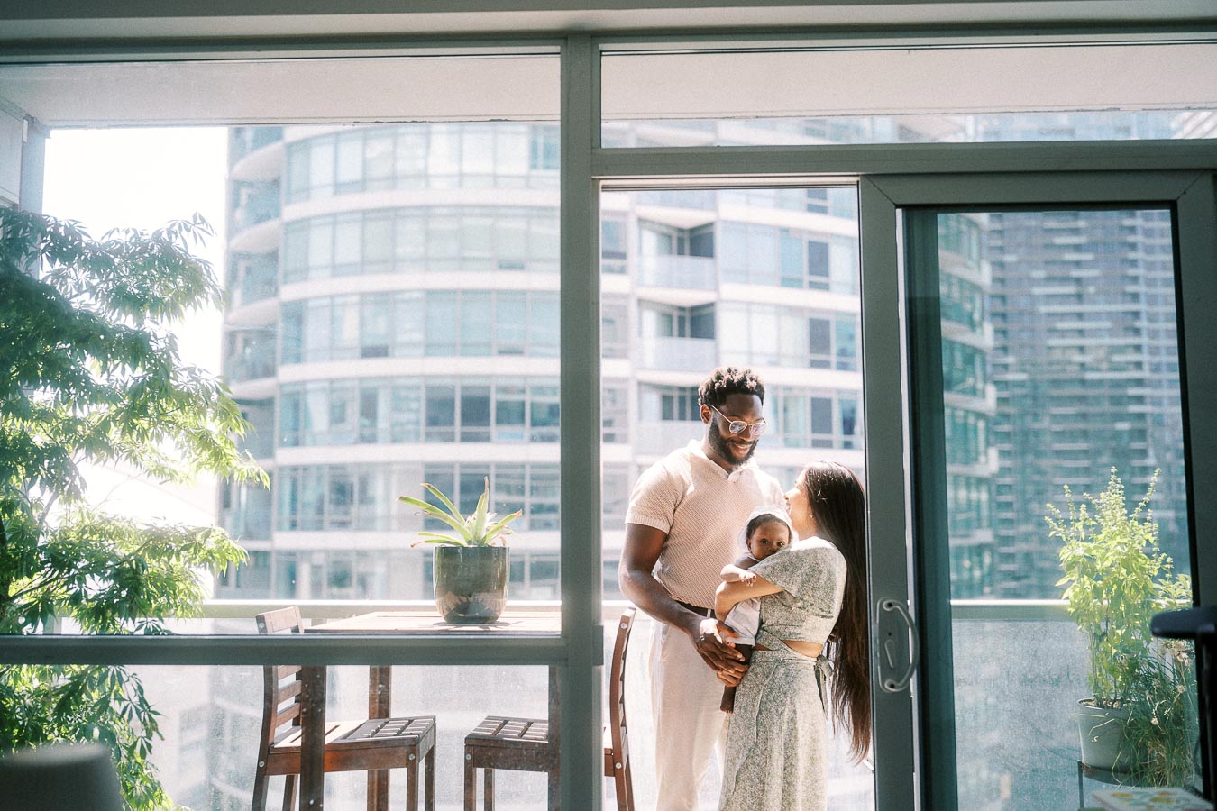 Parents holding their newborn on a bright Toronto balcony framed by glass doors