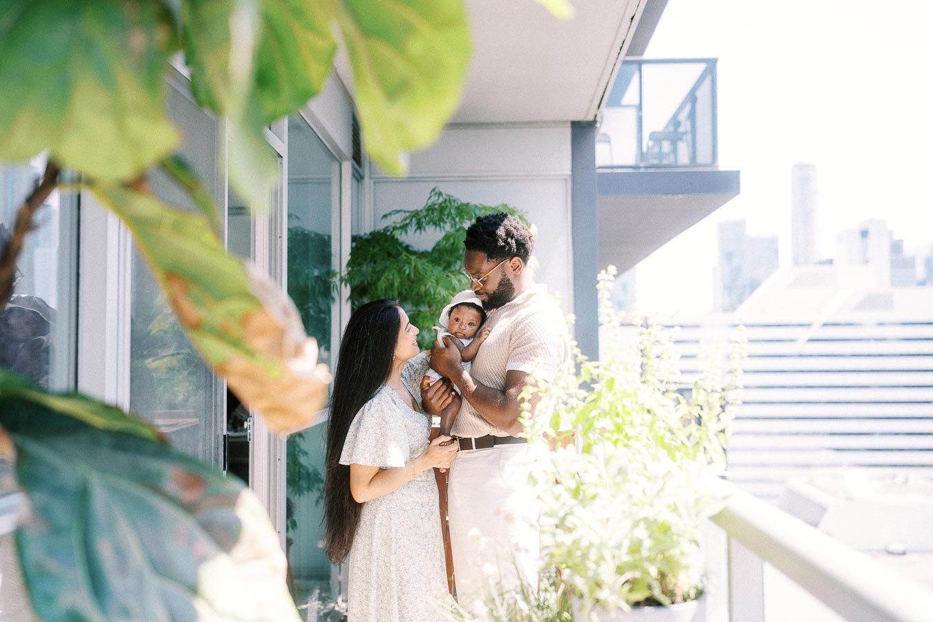 Parents holding their newborn daughter on a bright Toronto balcony surrounded by leafy plants.