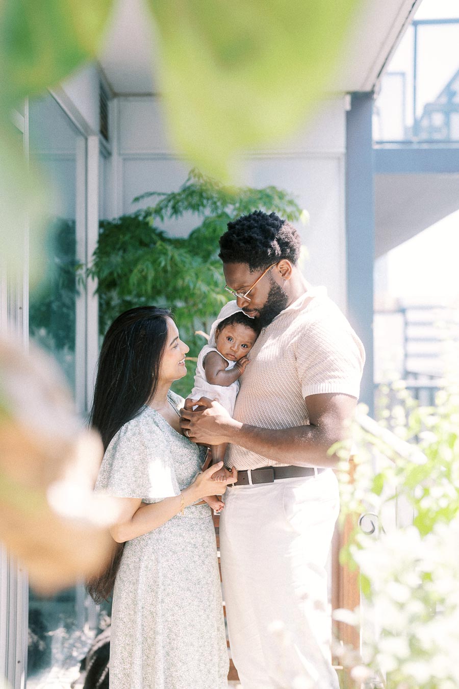Parents holding their newborn daughter on a sunny Toronto balcony framed by greenery.
