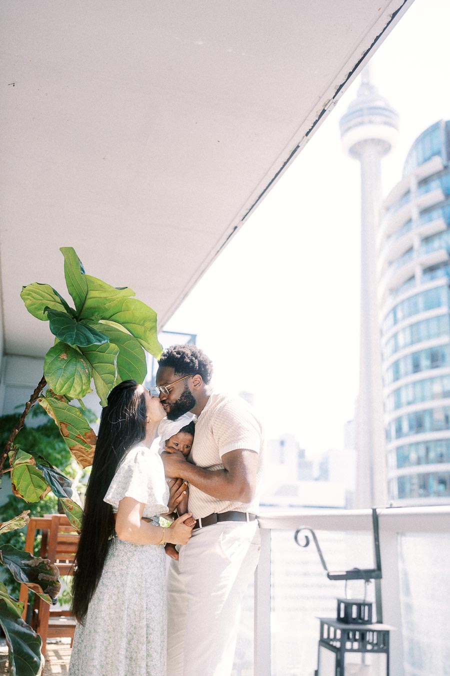 Parents kiss while holding newborn Isabella on a Toronto balcony with the CN Tower behind them.