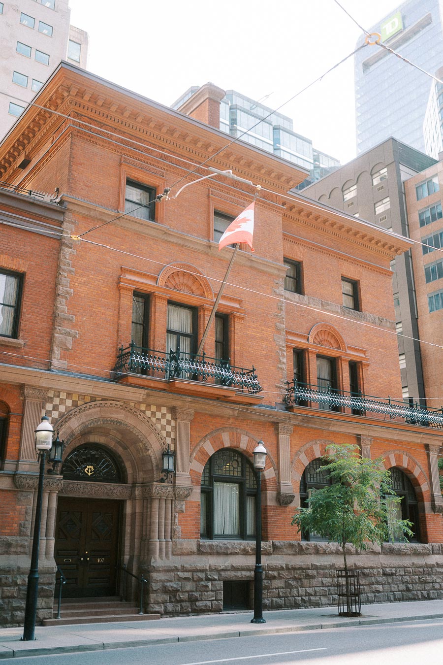 Historic red-brick Toronto Club facade with arched entrance, balconies, Canadian flag, and downtown towers.