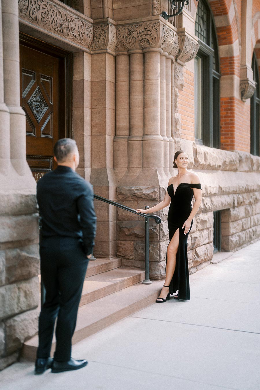Engaged couple in formal black attire posing by an ornate stone building entrance in Toronto.