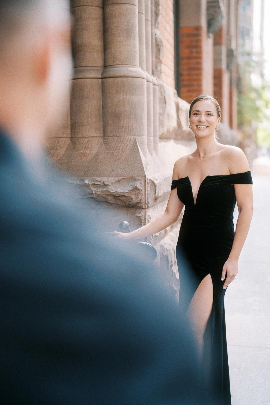 Woman in a black off-shoulder gown smiles beside a stone building, with partner blurred in foreground.