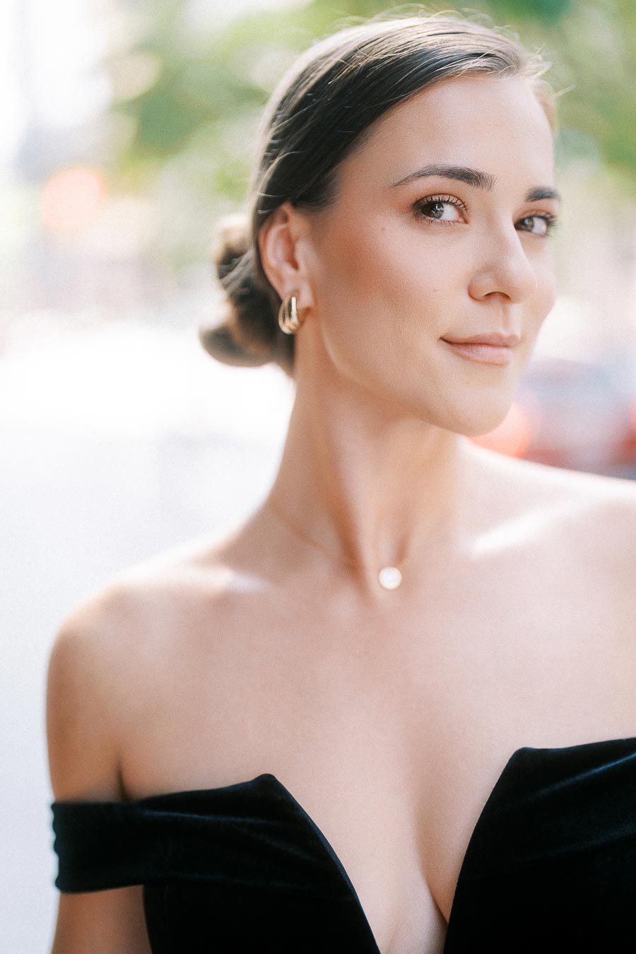 Bride-to-be in black off-shoulder dress with low bun and gold earrings, softly lit outdoors