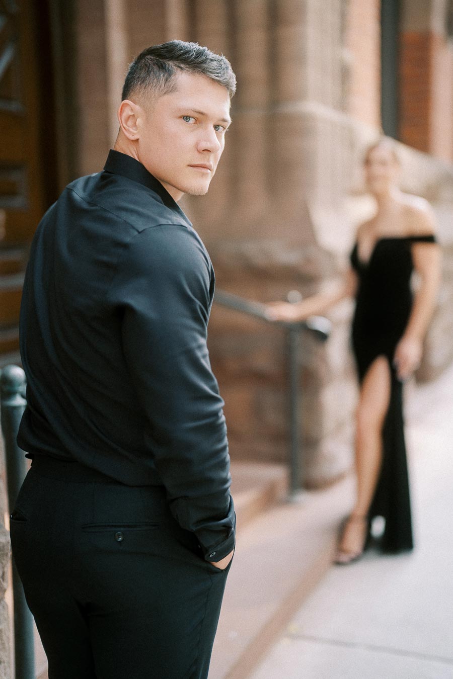 Man in black shirt looks back while woman in black dress stands blurred near stone columns.
