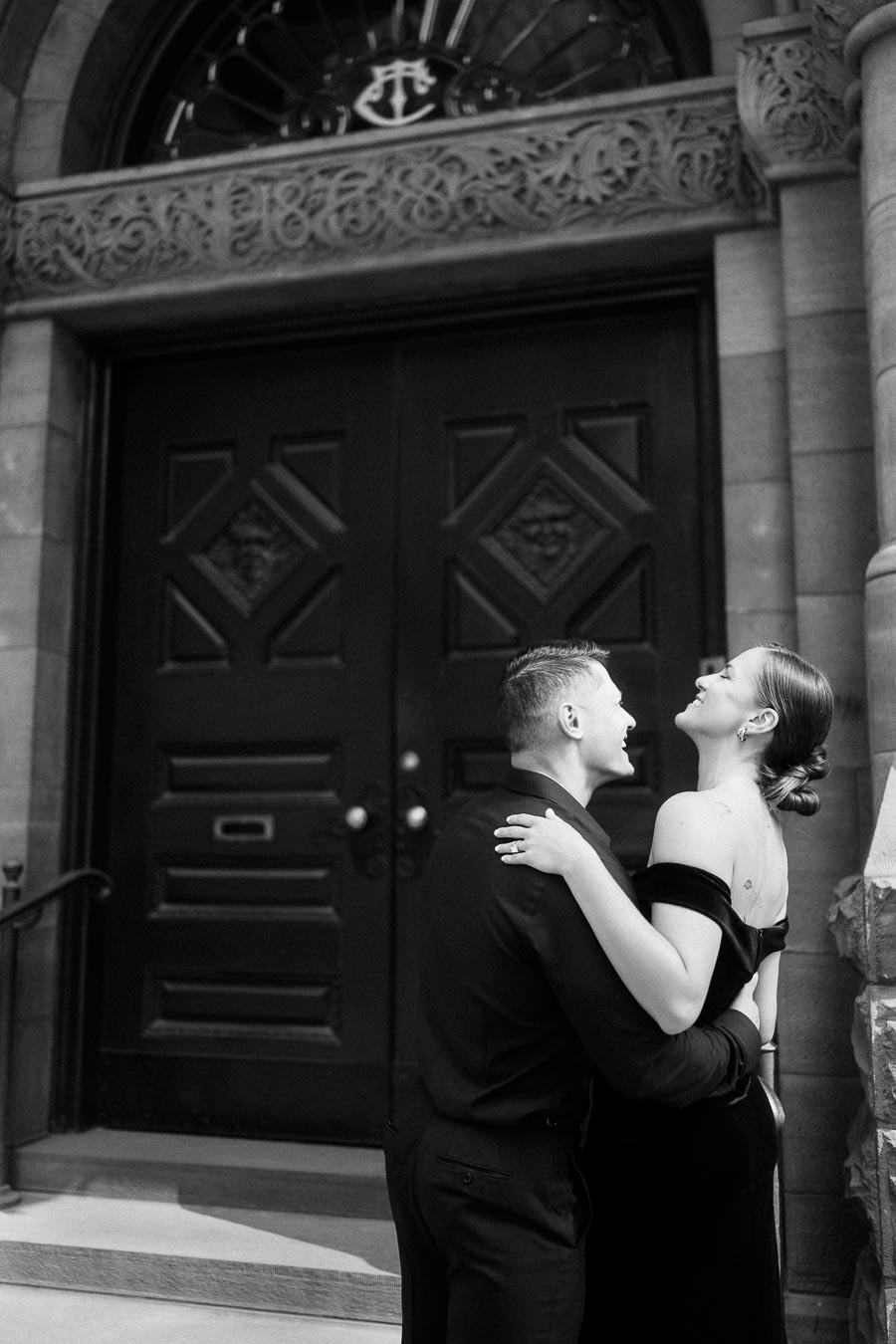 Couple laughing together in formal black outfits before ornate arched doors in downtown Toronto.