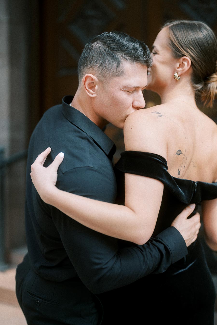 Groom-to-be kisses fiancée’s shoulder as they embrace in elegant black outfits in Toronto.