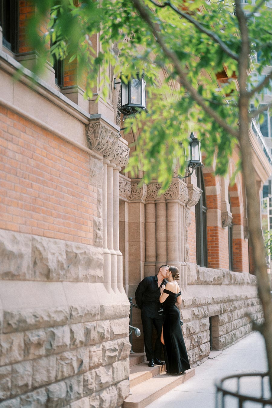 Couple kissing on stone steps beside a historic brick building framed by leafy branches.