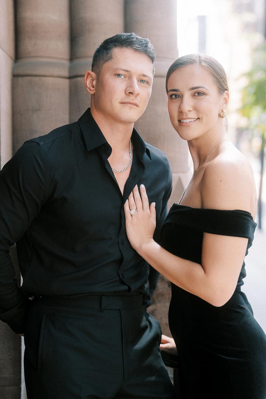 Engaged couple in black formal outfits posing beside stone columns, with her ring hand on his chest.