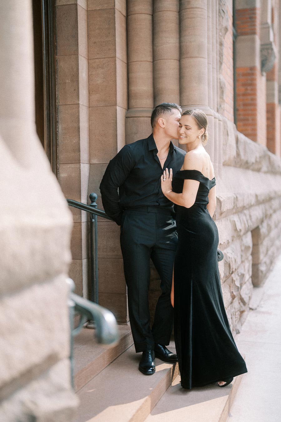 Couple in elegant black outfits embracing on stone steps beside a historic Toronto building