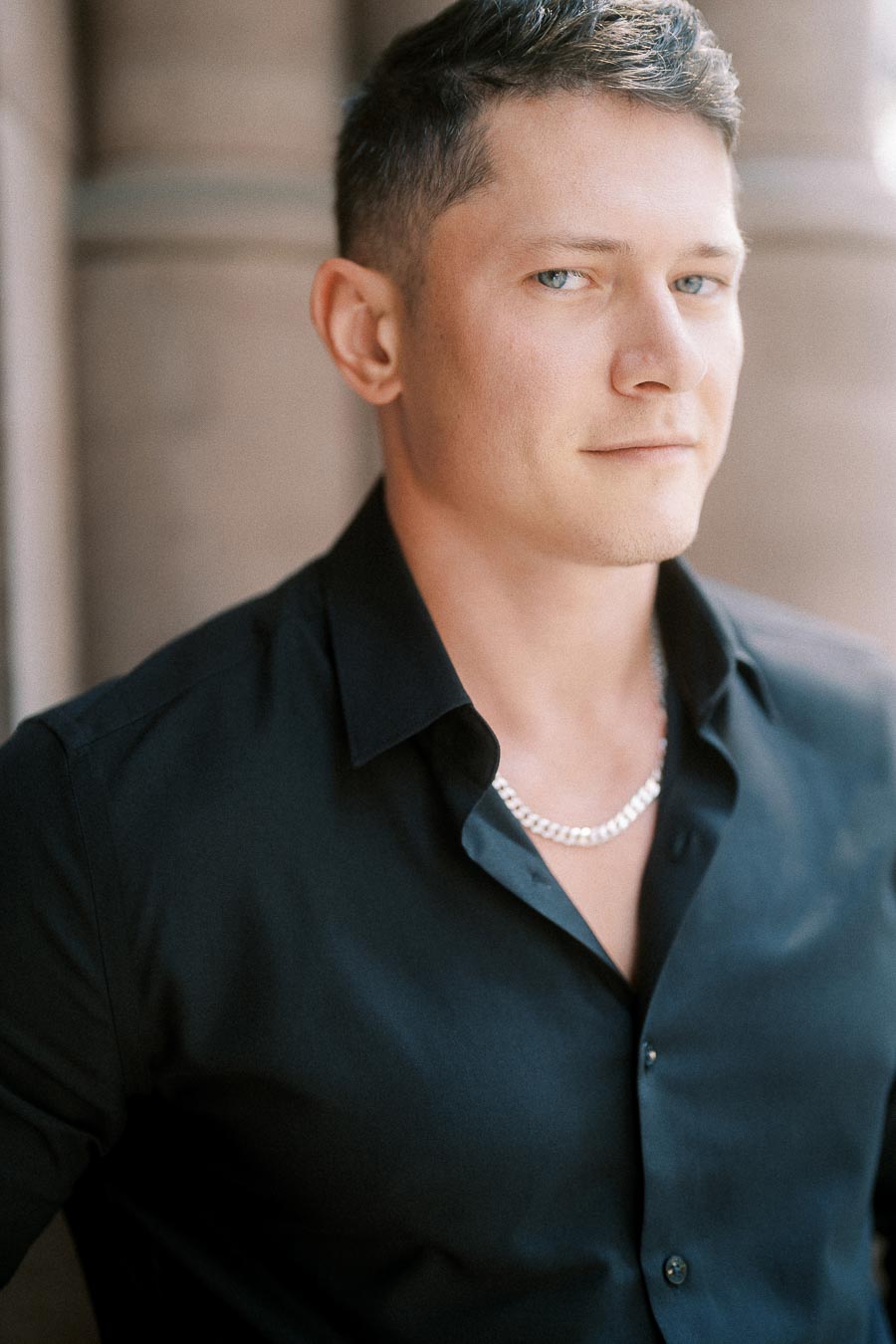 Close portrait of Alex in a black shirt and silver chain against soft stone architecture.