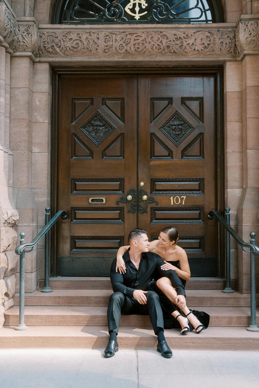 Couple in black outfits sitting on stone steps before ornate wooden doors numbered 107 in Toronto.