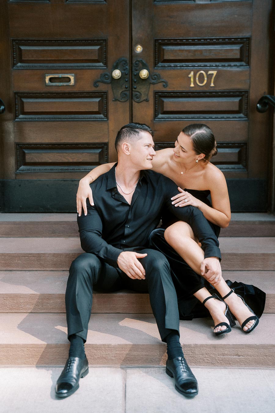 Engaged couple in black outfits sitting on stone steps before ornate wooden doors numbered 107.
