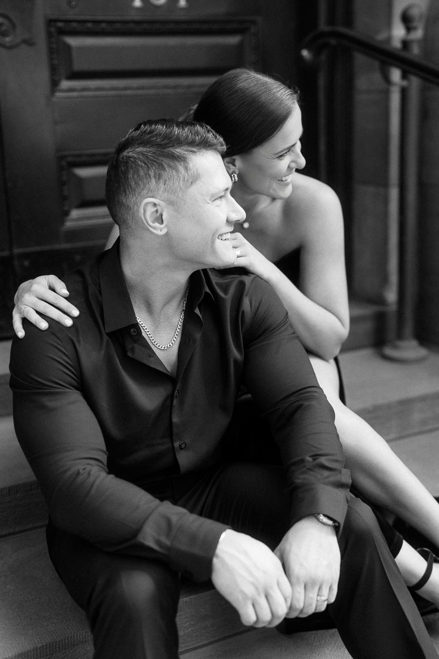 Couple sitting close on stone steps, smiling together beside a dark doorway and railing.