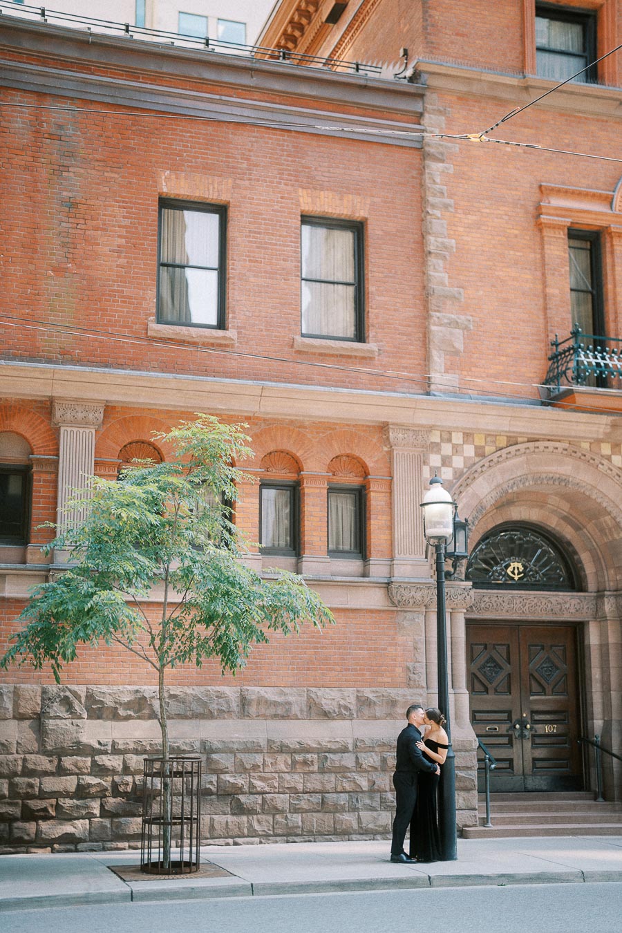 Couple embraces on a downtown Toronto sidewalk beside a red brick heritage building and arched doorway.