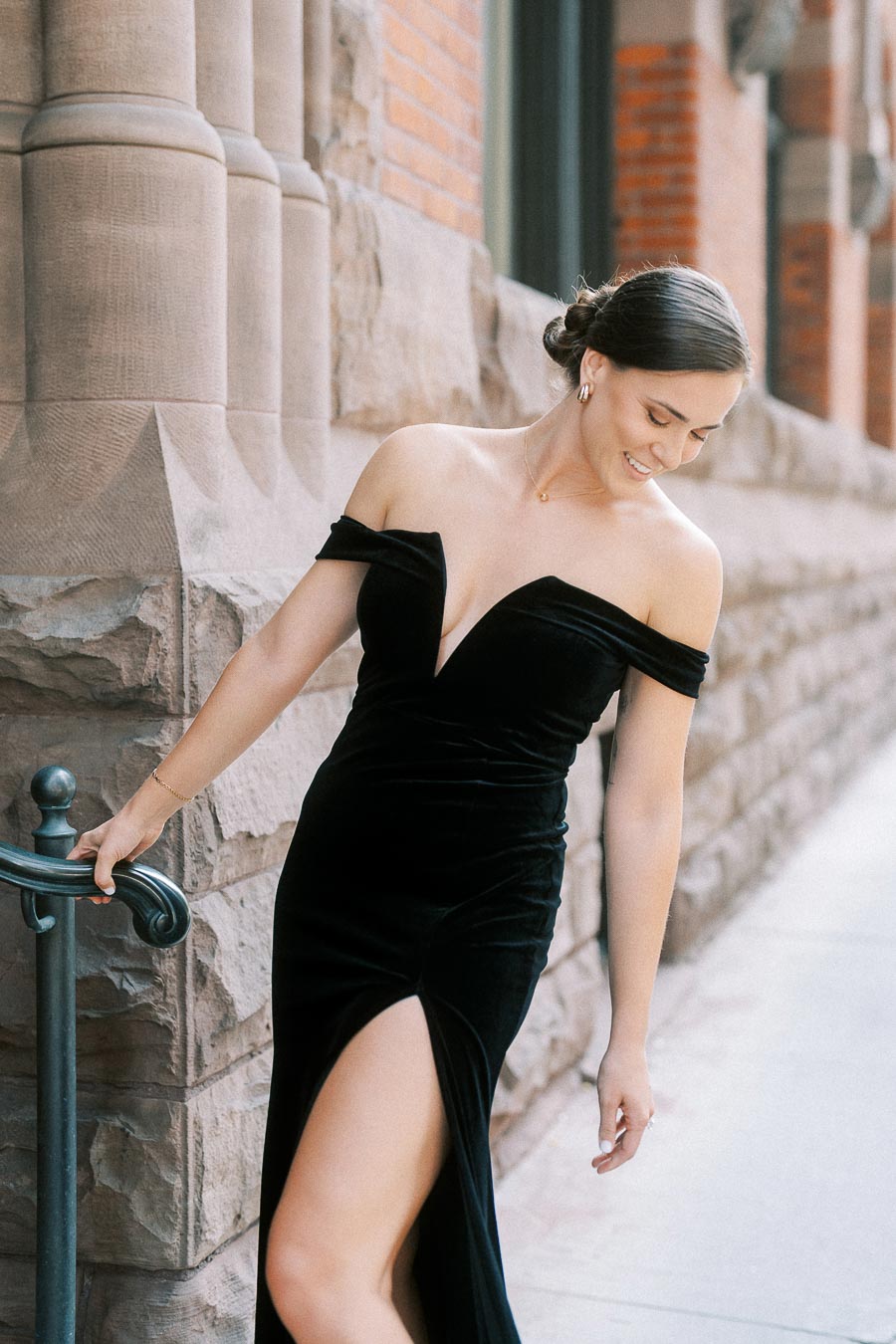 Woman in a black off-shoulder gown holds a railing beside a stone and brick Toronto building.