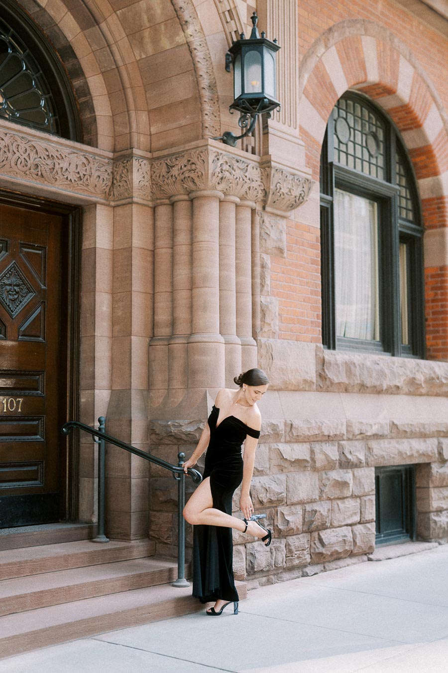 Woman in black gown adjusts her heel beside an ornate stone entrance in downtown Toronto.