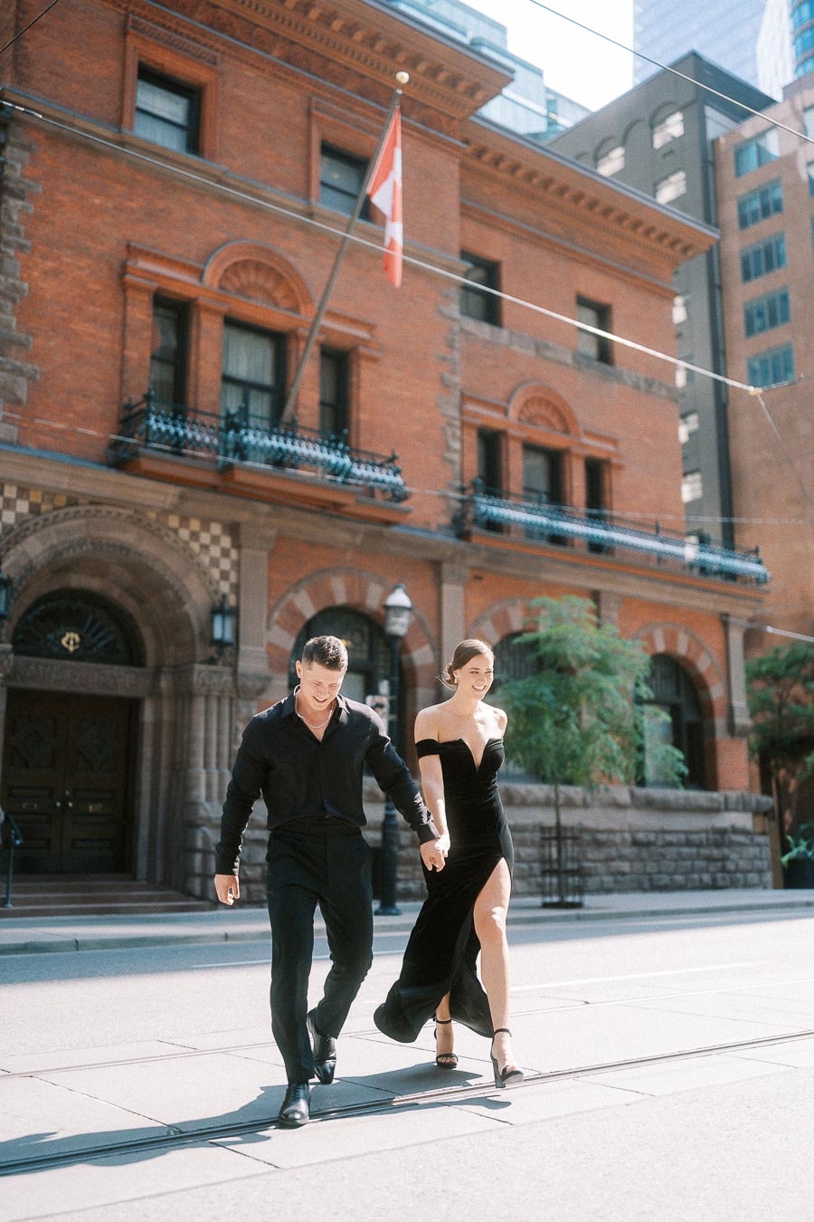 Couple in black formalwear walking hand in hand past a red brick downtown Toronto building.