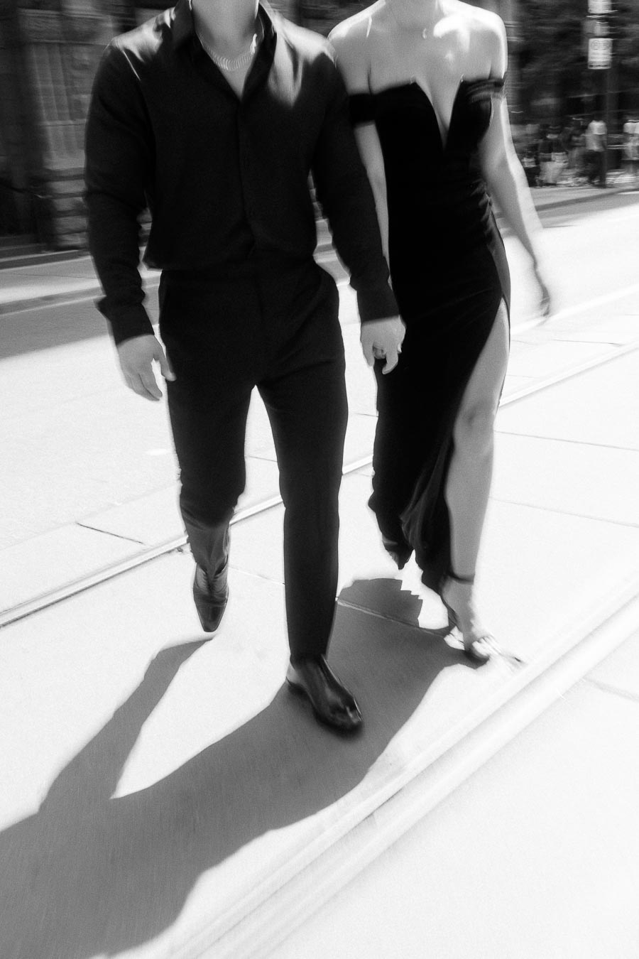 Couple in formal black outfits walking hand in hand along a sunny downtown Toronto street