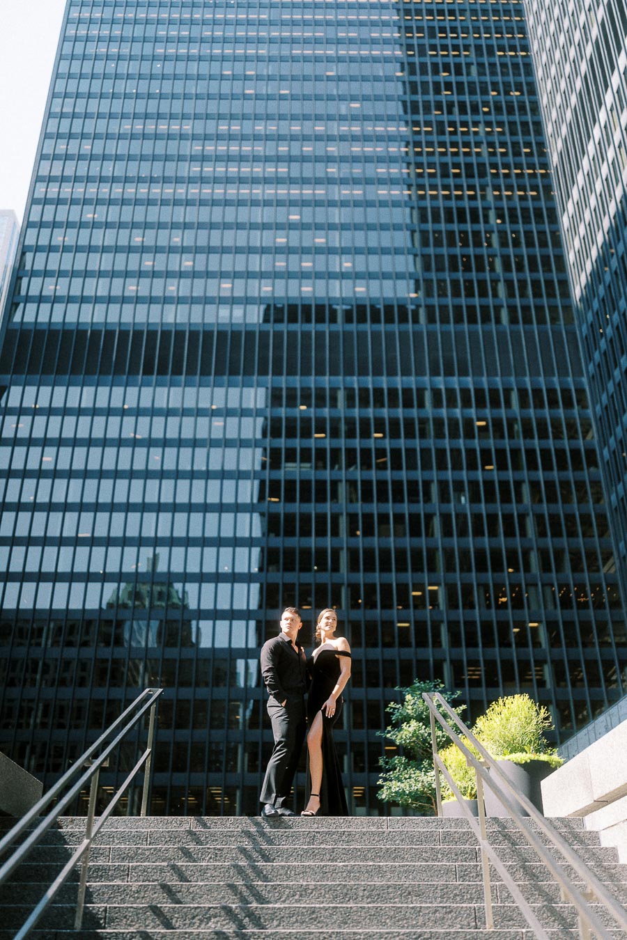 Couple in black formalwear posing on steps before tall glass towers in downtown Toronto.