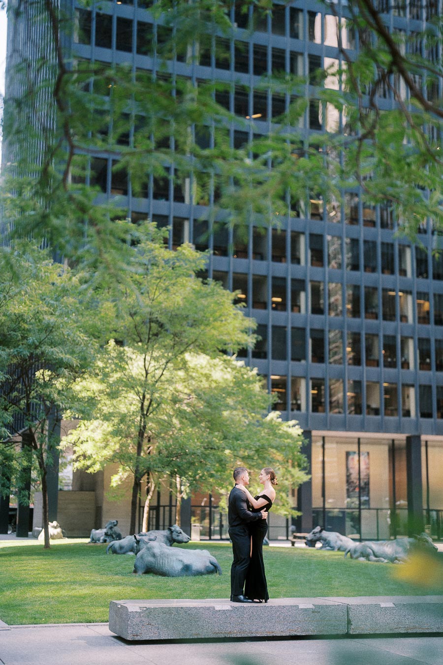 Couple in formal black attire embracing on a downtown Toronto lawn with sculptural cows nearby