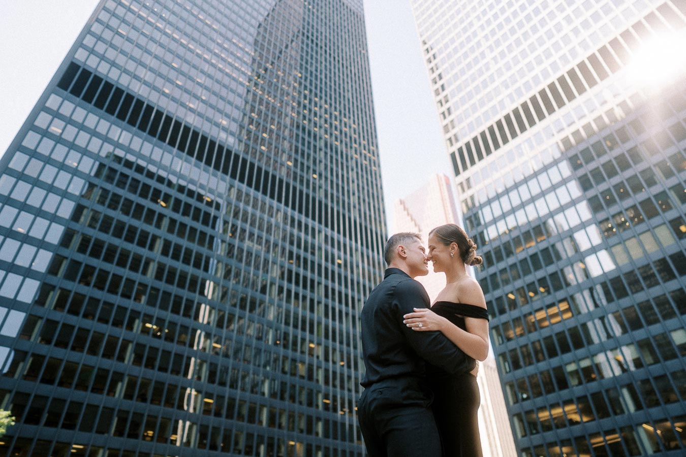 Couple embracing beneath tall glass office towers in downtown Toronto with sunlight flaring above.