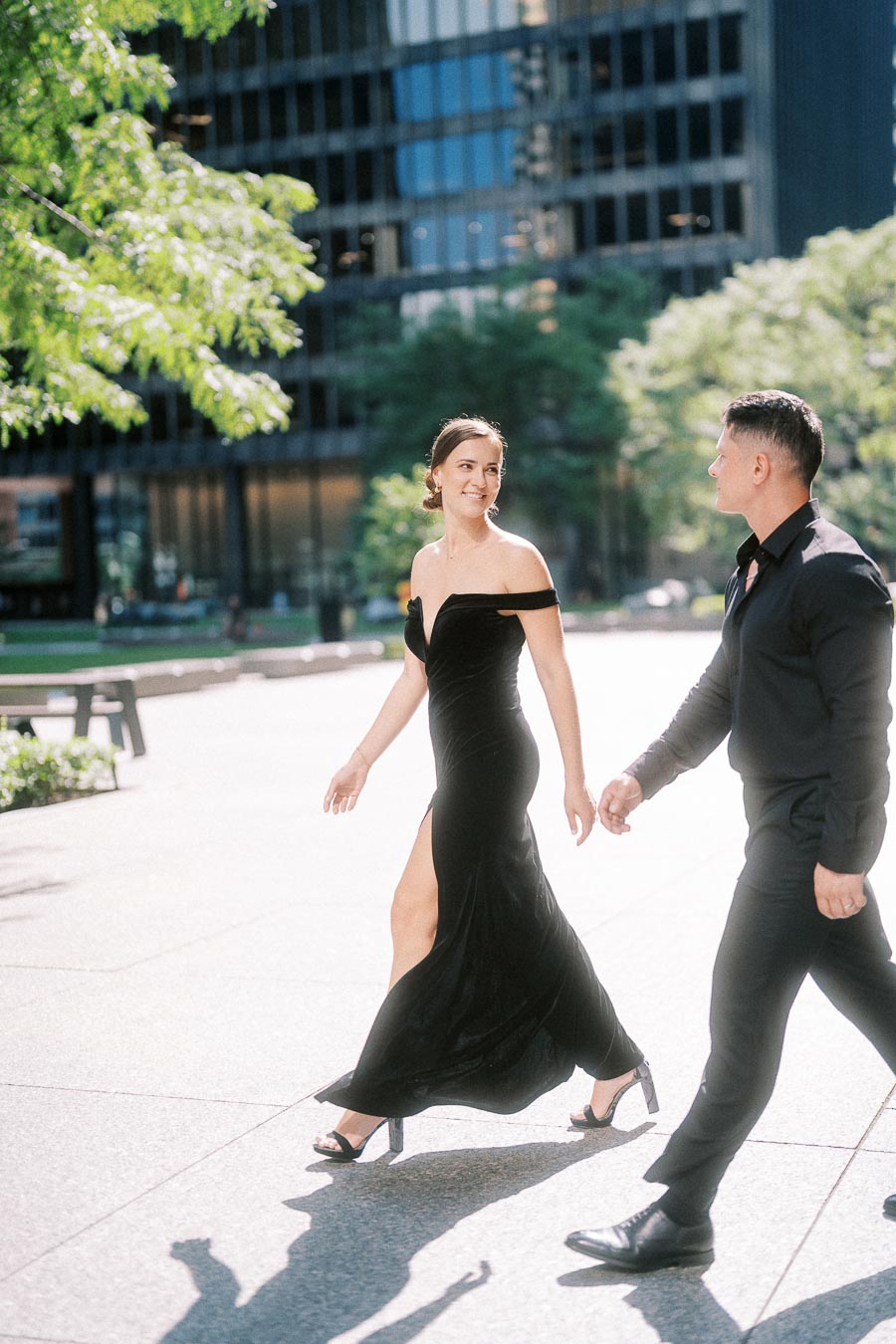 Couple in black formalwear walks hand in hand through a sunny downtown Toronto plaza.