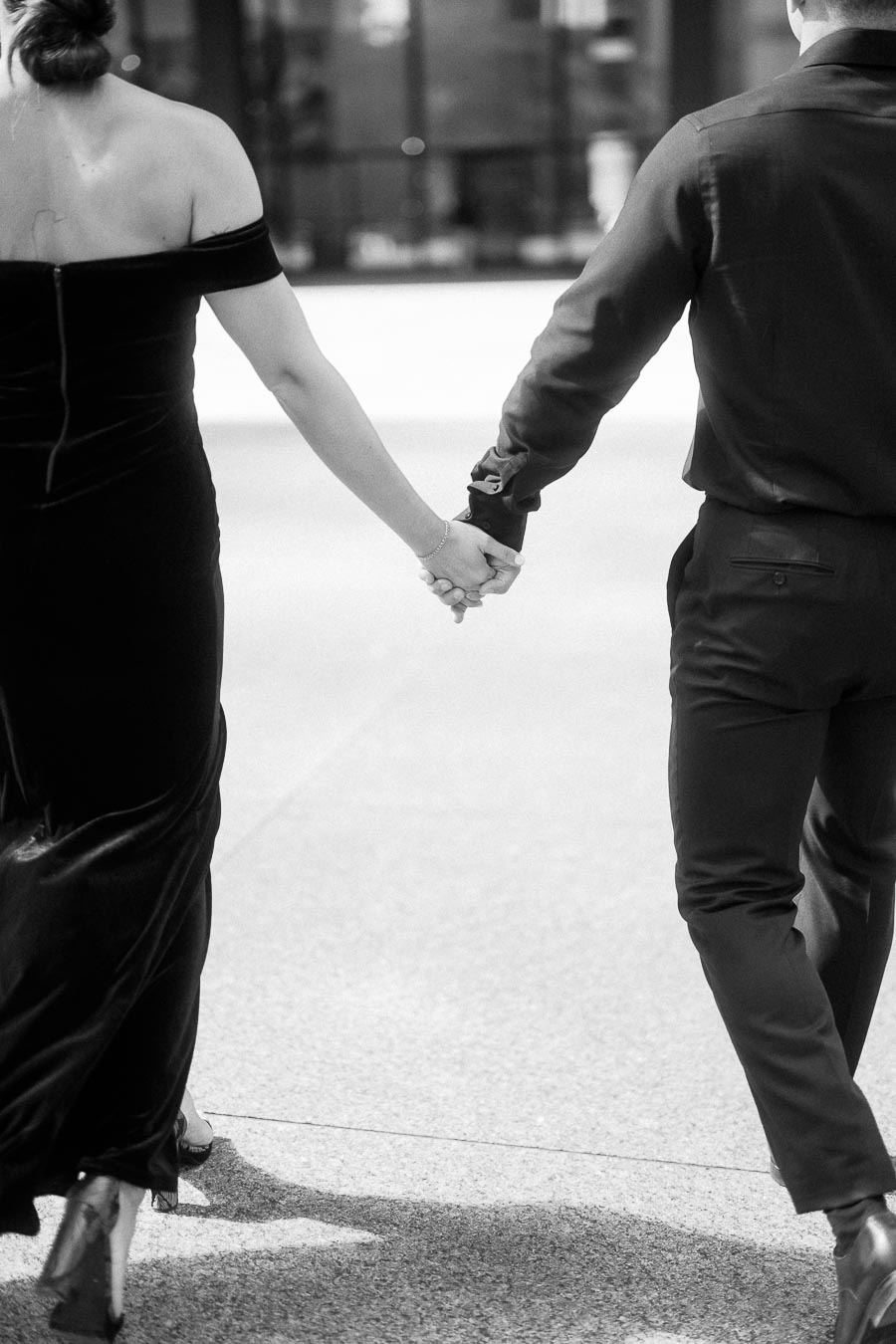 Couple in formal black attire walking hand in hand along a sunlit urban sidewalk.