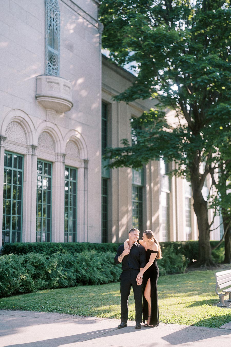 Engaged couple in black outfits standing beside a historic building and leafy trees in Toronto.