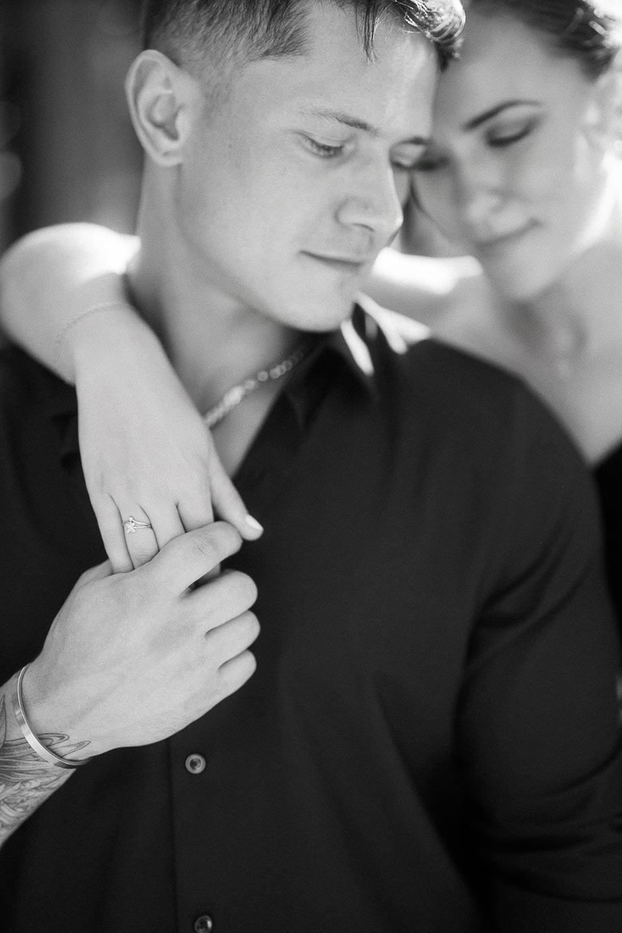 Black-and-white close-up of an engaged couple, with her ringed hand resting on his shoulder.