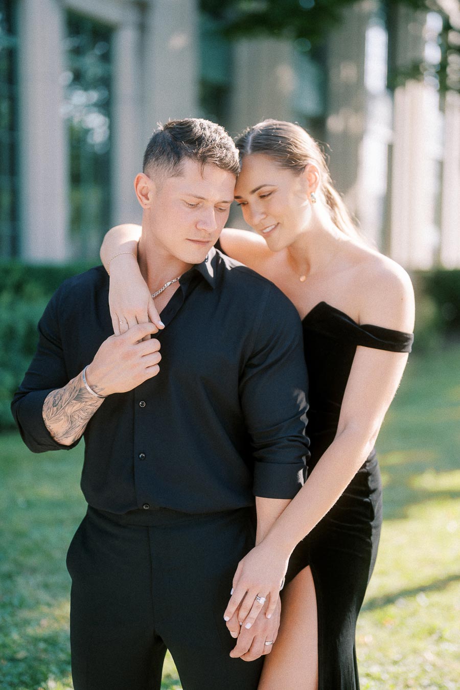 Couple in black formal outfits embracing outdoors beside sunlit columns and grass in Toronto.
