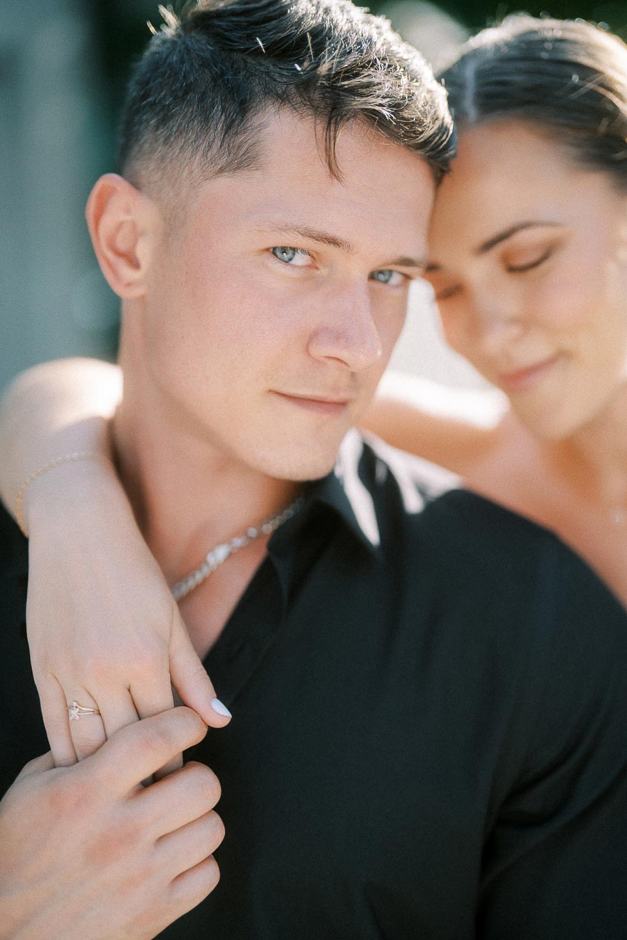 Close portrait of engaged couple with woman's ring hand resting on man's shoulder in soft sunlight.