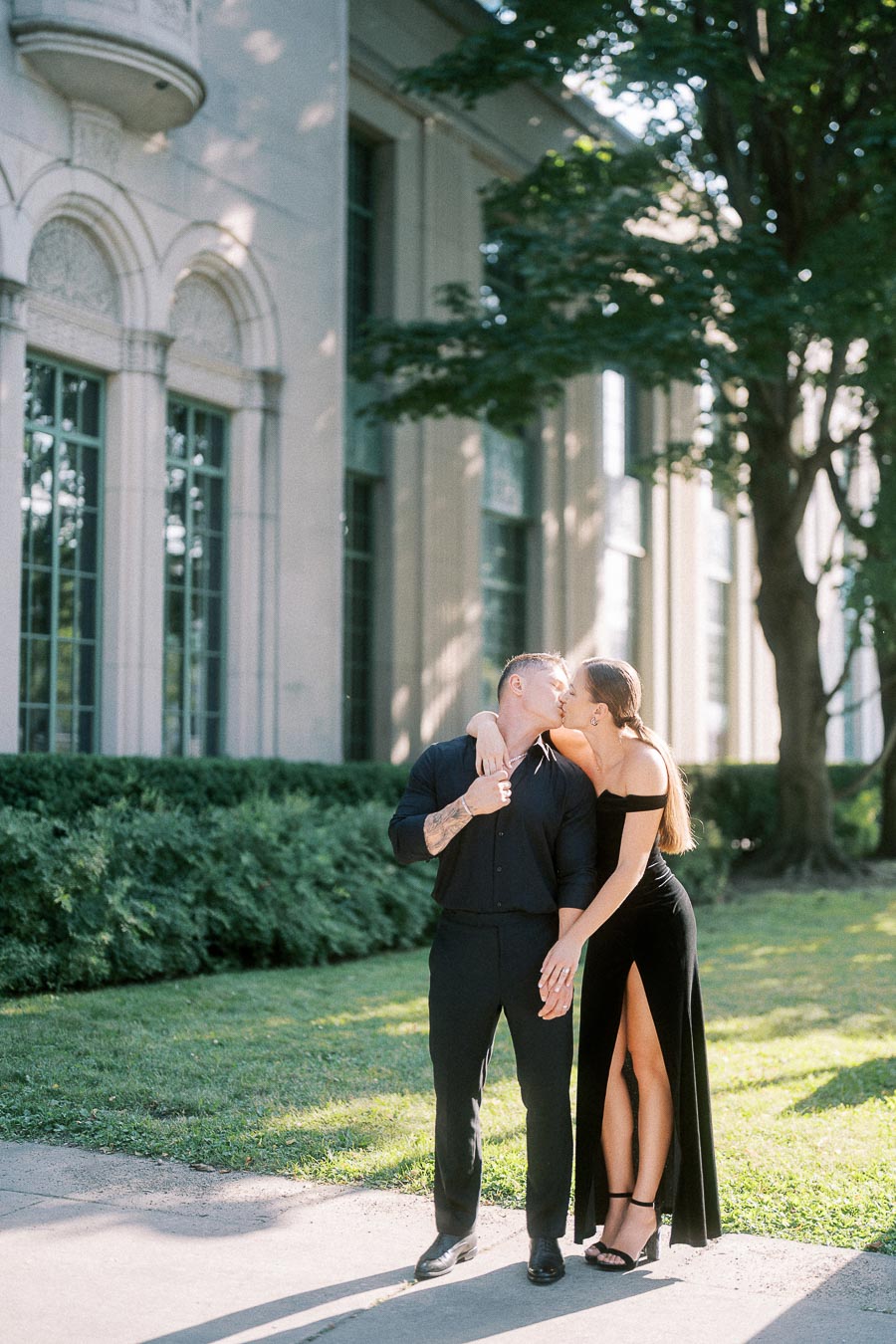 Couple in black outfits kissing beside an elegant stone building and sunlit trees in Toronto.