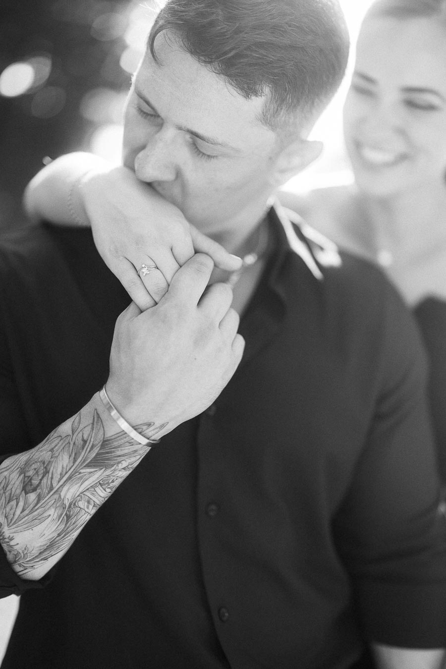 Black-and-white close-up of groom kissing bride’s ringed hand as she smiles behind him.