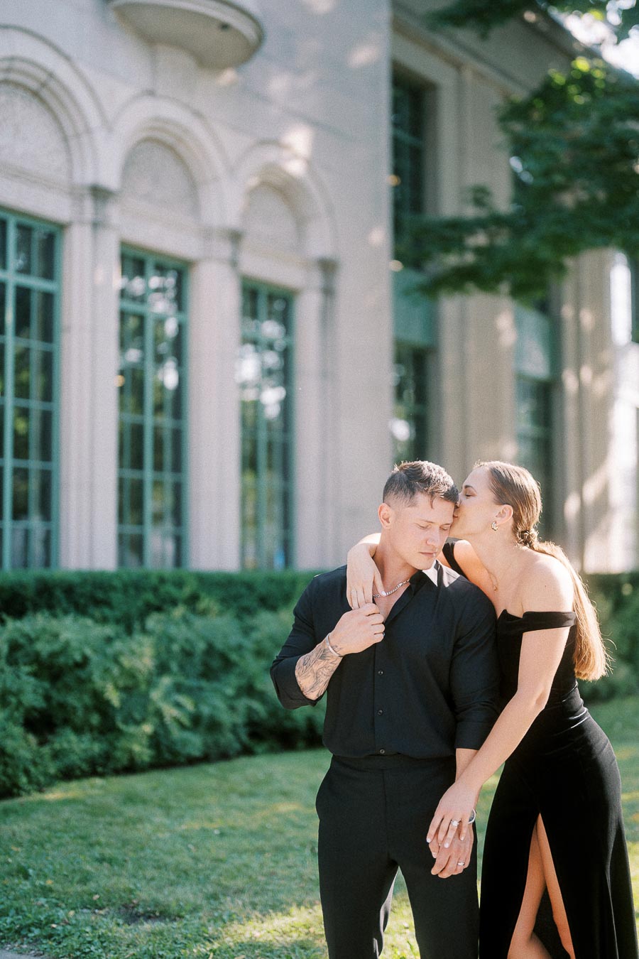 Couple in black formal outfits holding hands beside an elegant stone building and green lawn.