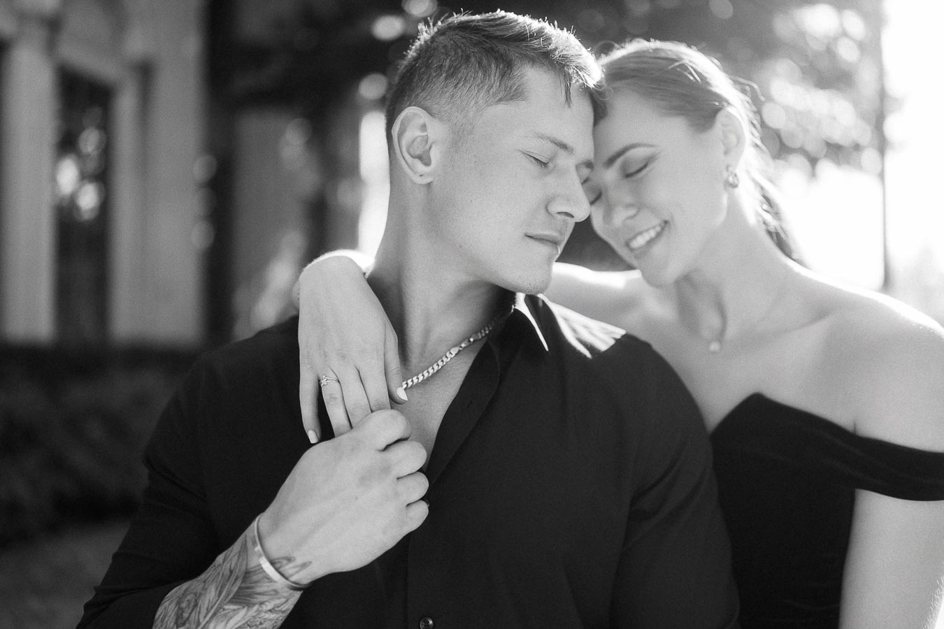 Black-and-white close portrait of an engaged couple embracing, her ring visible on his shoulder.