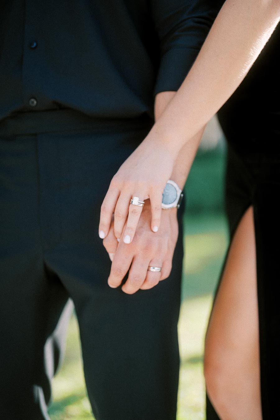 Close-up of a couple holding hands, showing engagement and wedding rings against black formalwear.