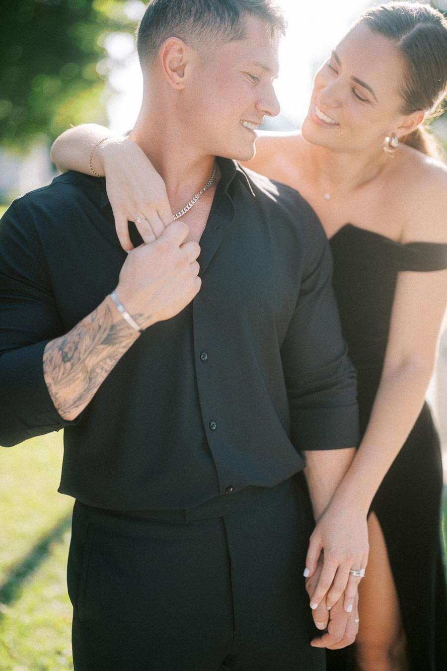Smiling couple in black outfits holding hands outdoors, with her arm draped over his shoulder.