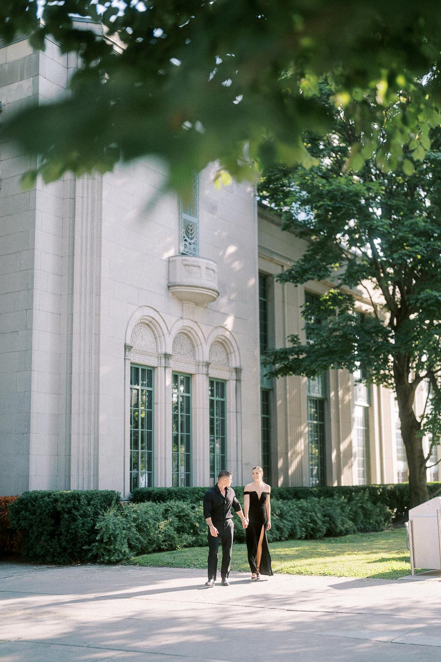 Engaged couple walking hand in hand outside an elegant stone building framed by leafy trees.