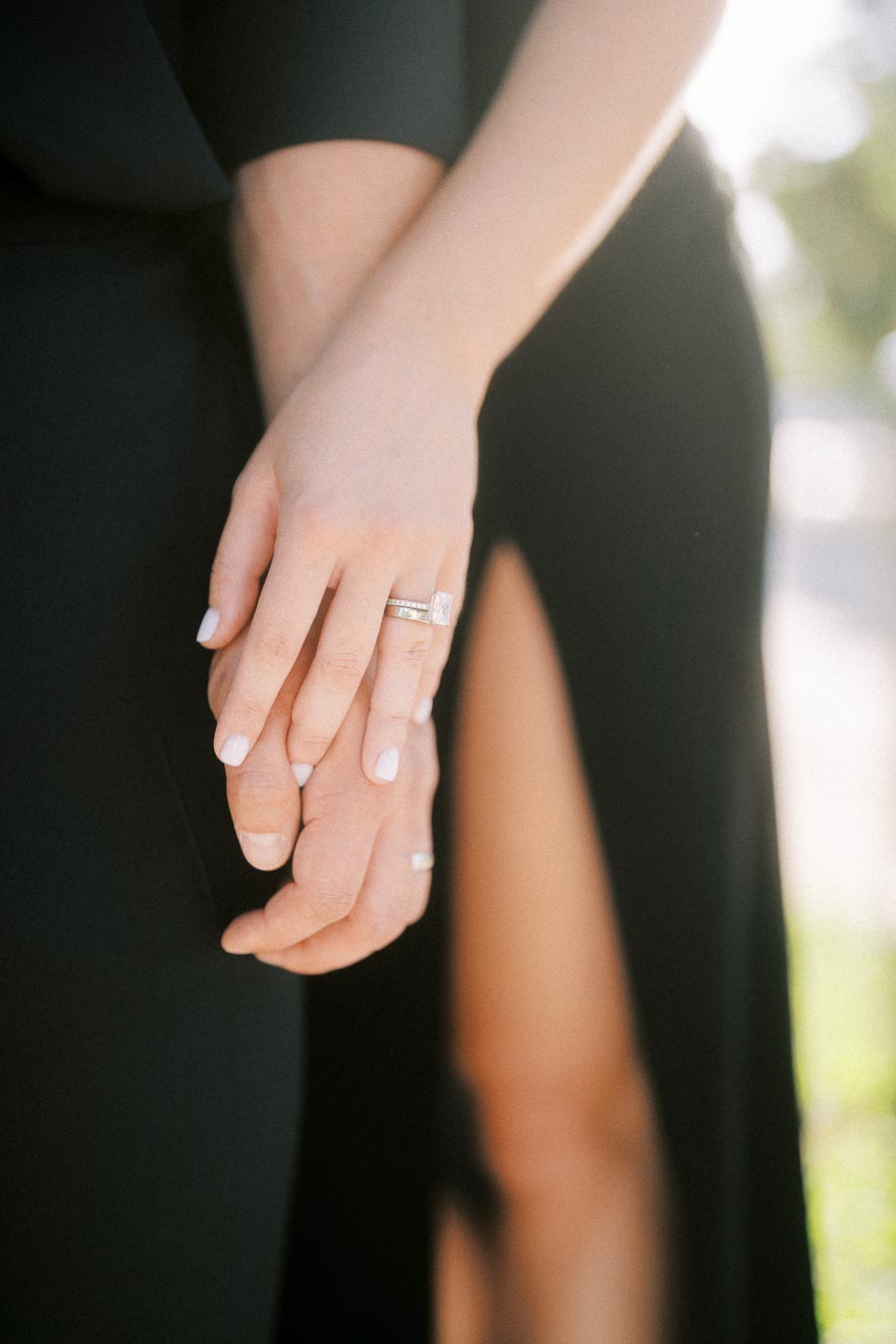 Close-up of a couple holding hands, highlighting a diamond engagement ring against black formal attire.