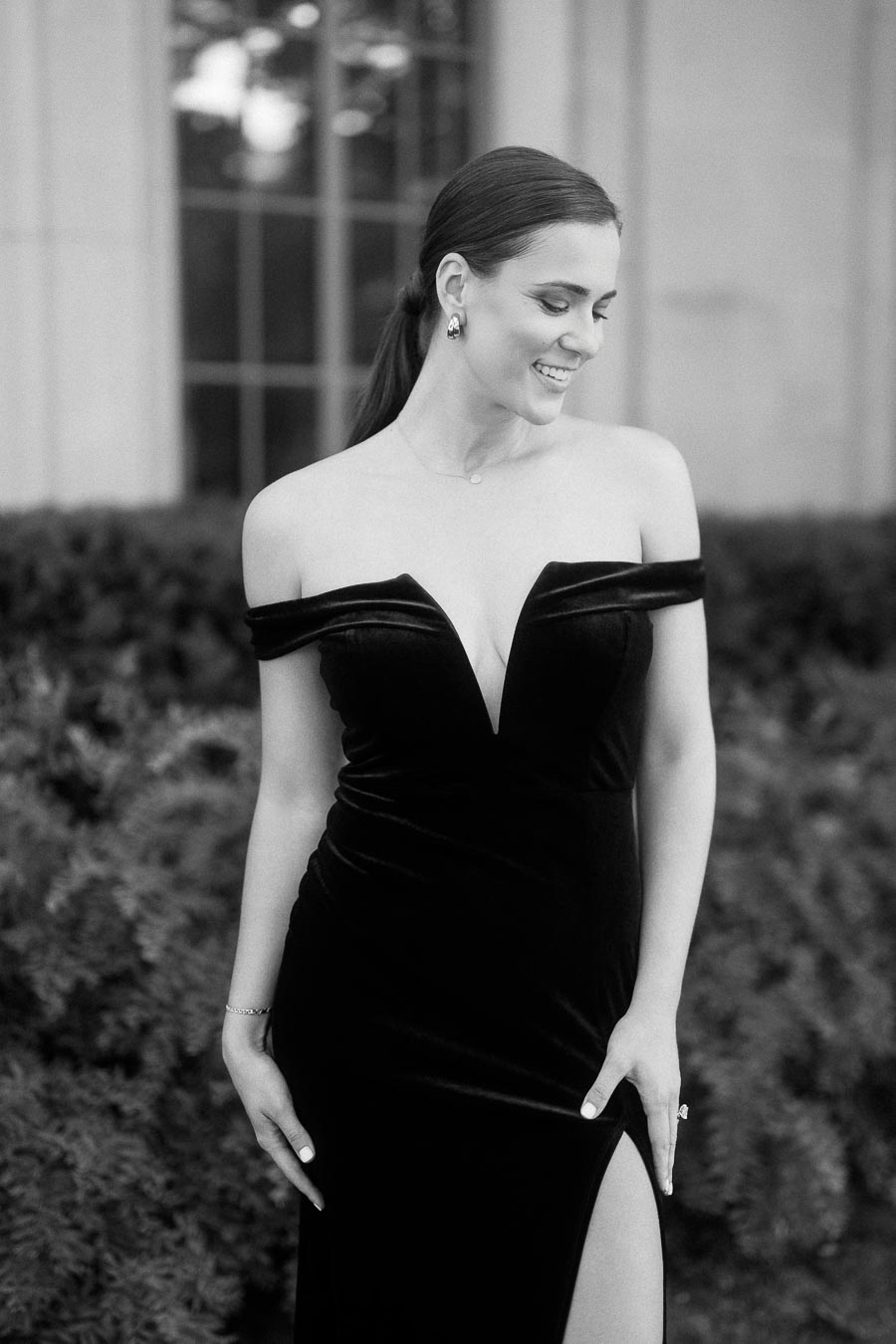 Woman in an off-shoulder black gown smiling beside hedges and tall windows.